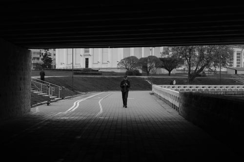 Free Black and white photo of an urban walkway with a man walking under a bridge, smartphone in hand. Stock Photo