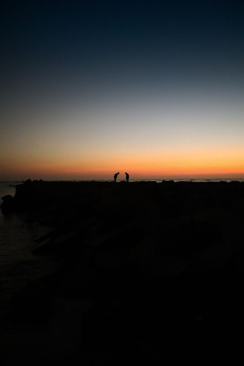 Free Two people silhouetted against a vibrant twilight sky on a rocky seacoast. Stock Photo