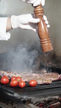 A chef seasons grilled meat and cherry tomatoes with fresh pepper on a smoky grill.
