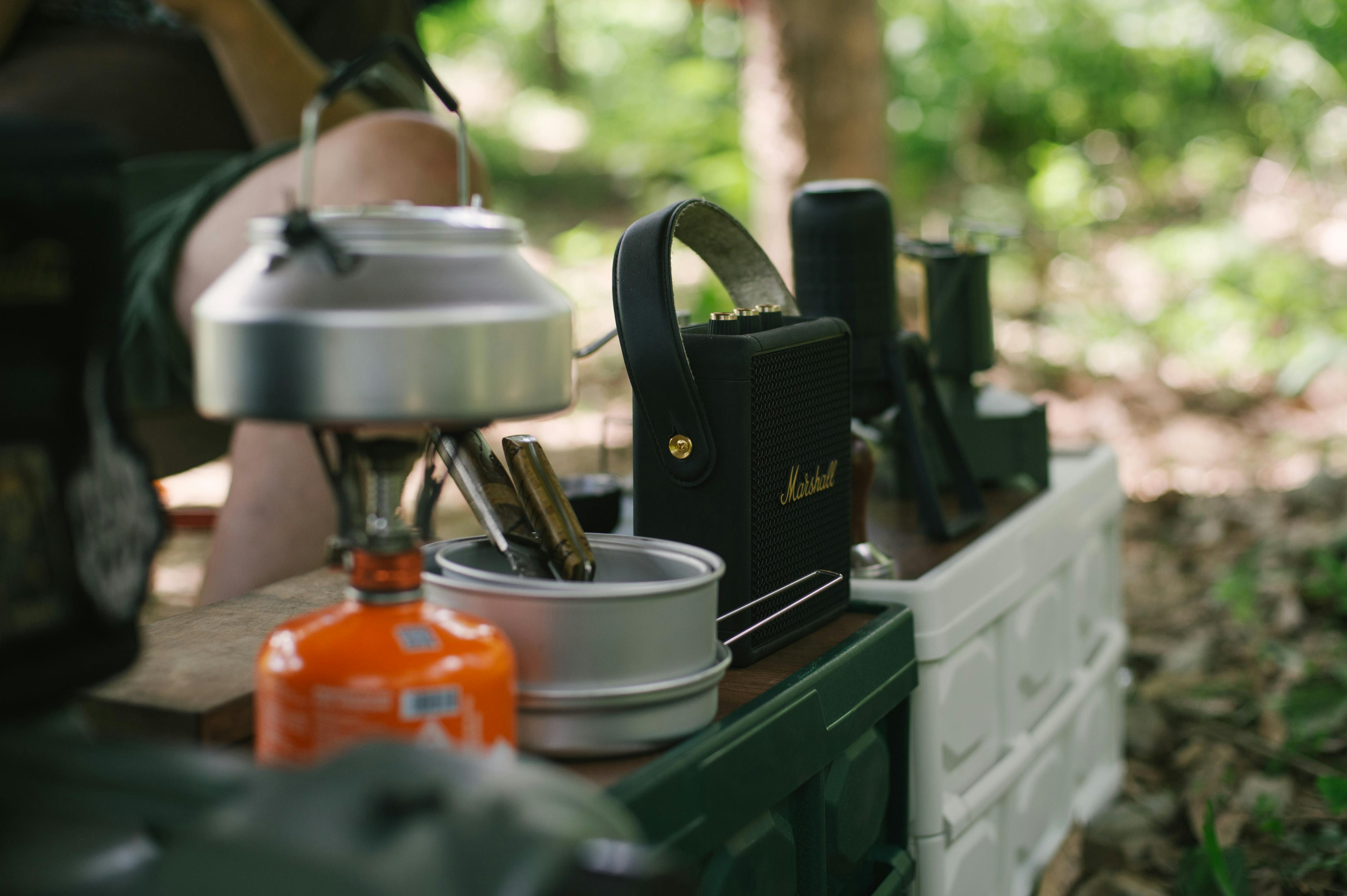 Outdoor camping gear including a gas stove and kettle on a table in a forest setting