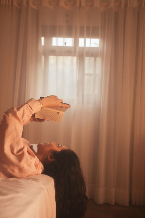 Free Woman lying in a sunlit room reading a book, enjoying a peaceful moment. Stock Photo