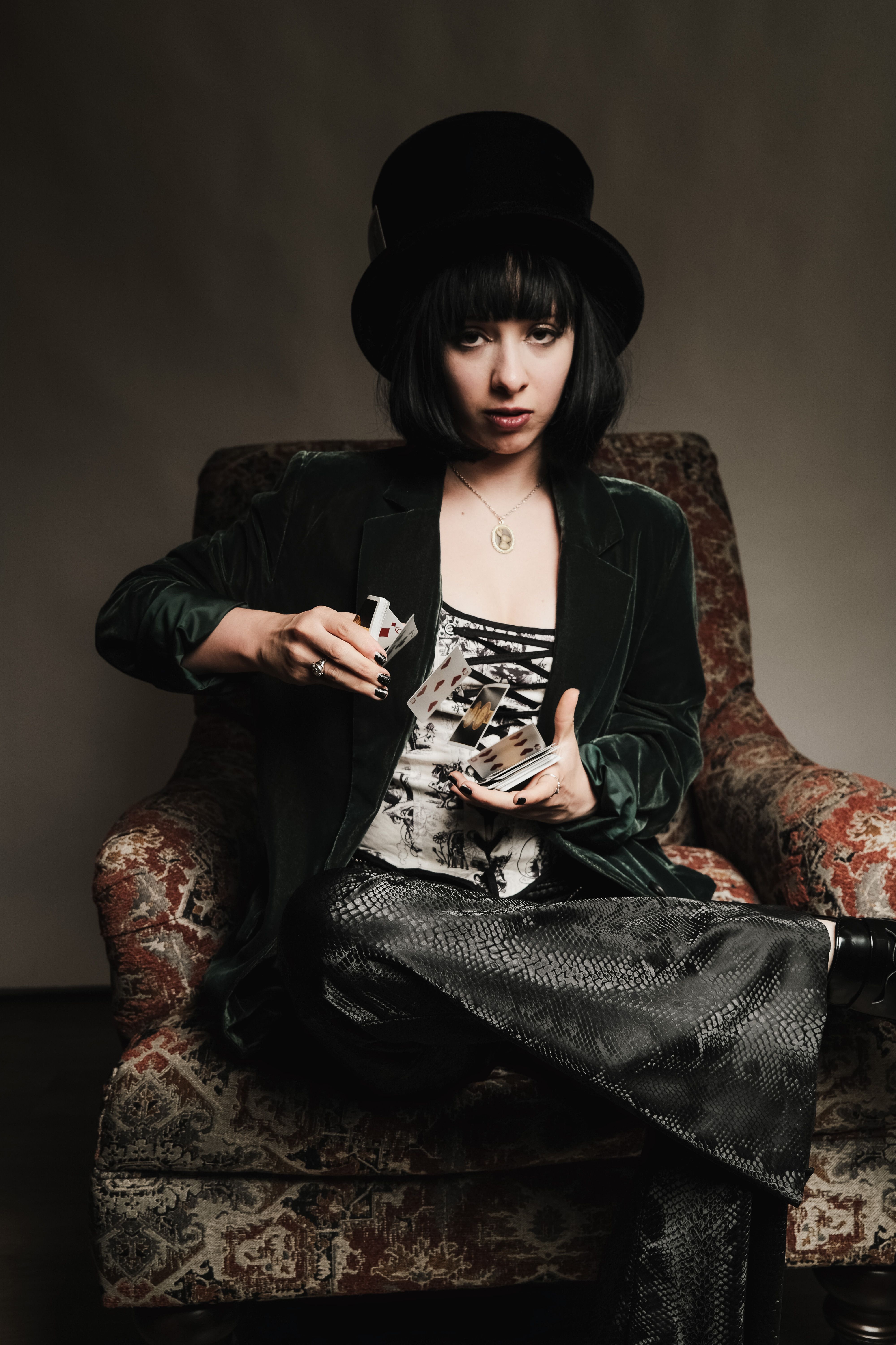 Woman in velvet jacket and top hat performing a card trick while seated indoors.