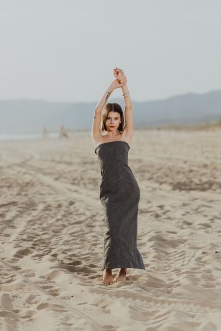 A woman in a grey dress poses elegantly on a sunlit beach with mountains in the background.
