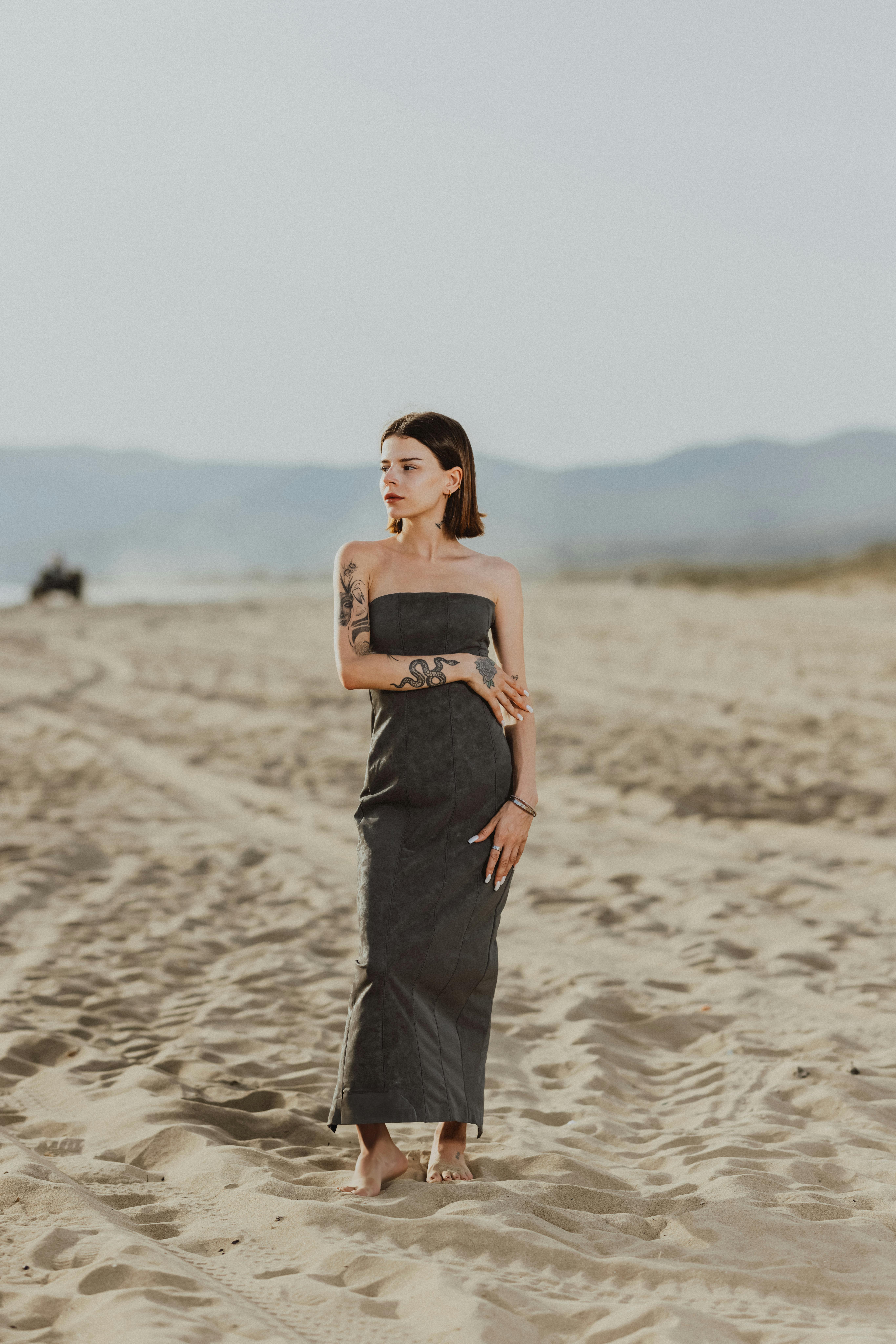 A woman in a grey dress stands elegantly on a sandy beach, under the sunlight.