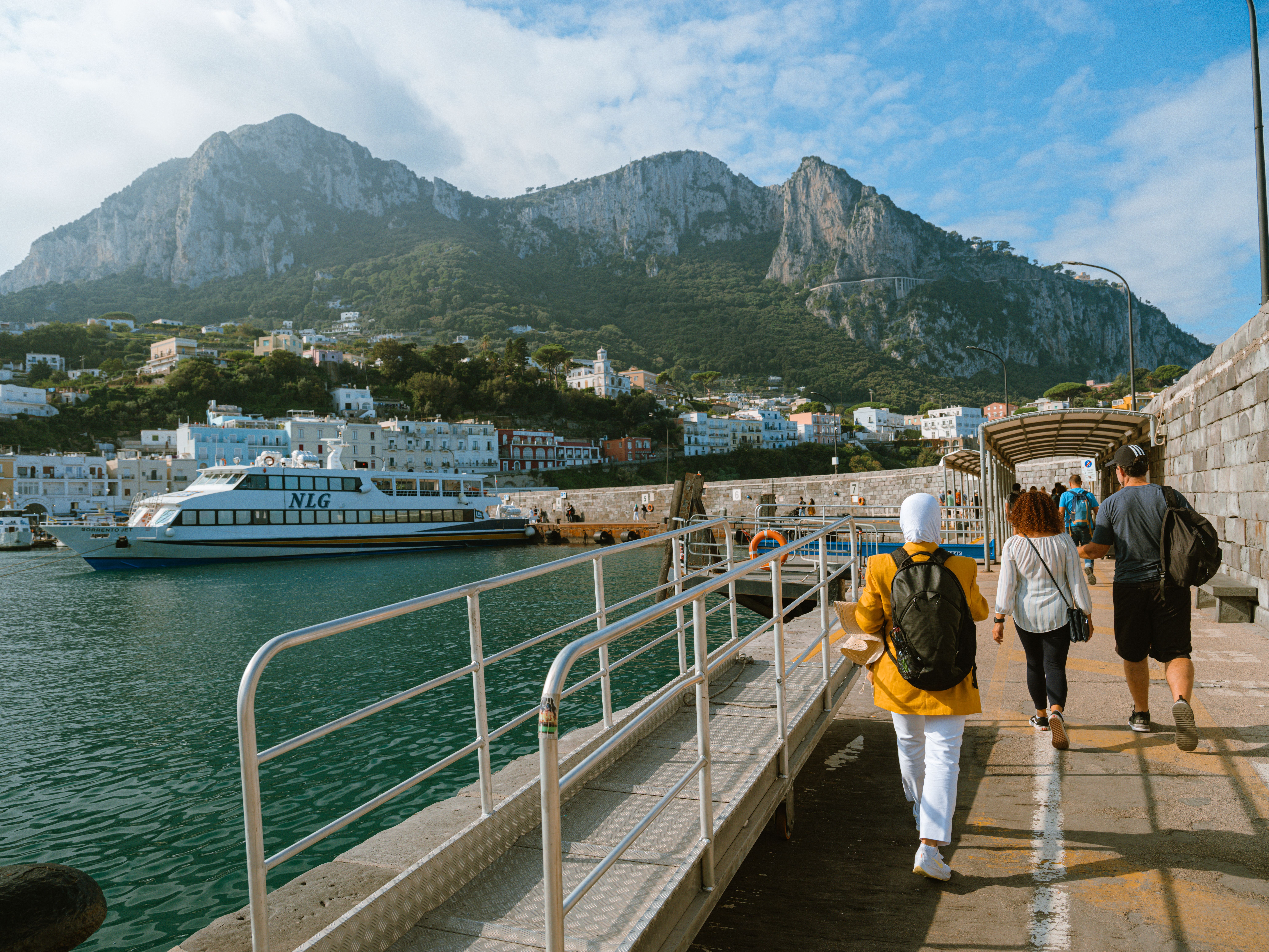 A picturesque view of tourists walking on the pier of Capri Island, Italy, with mountains and sea.