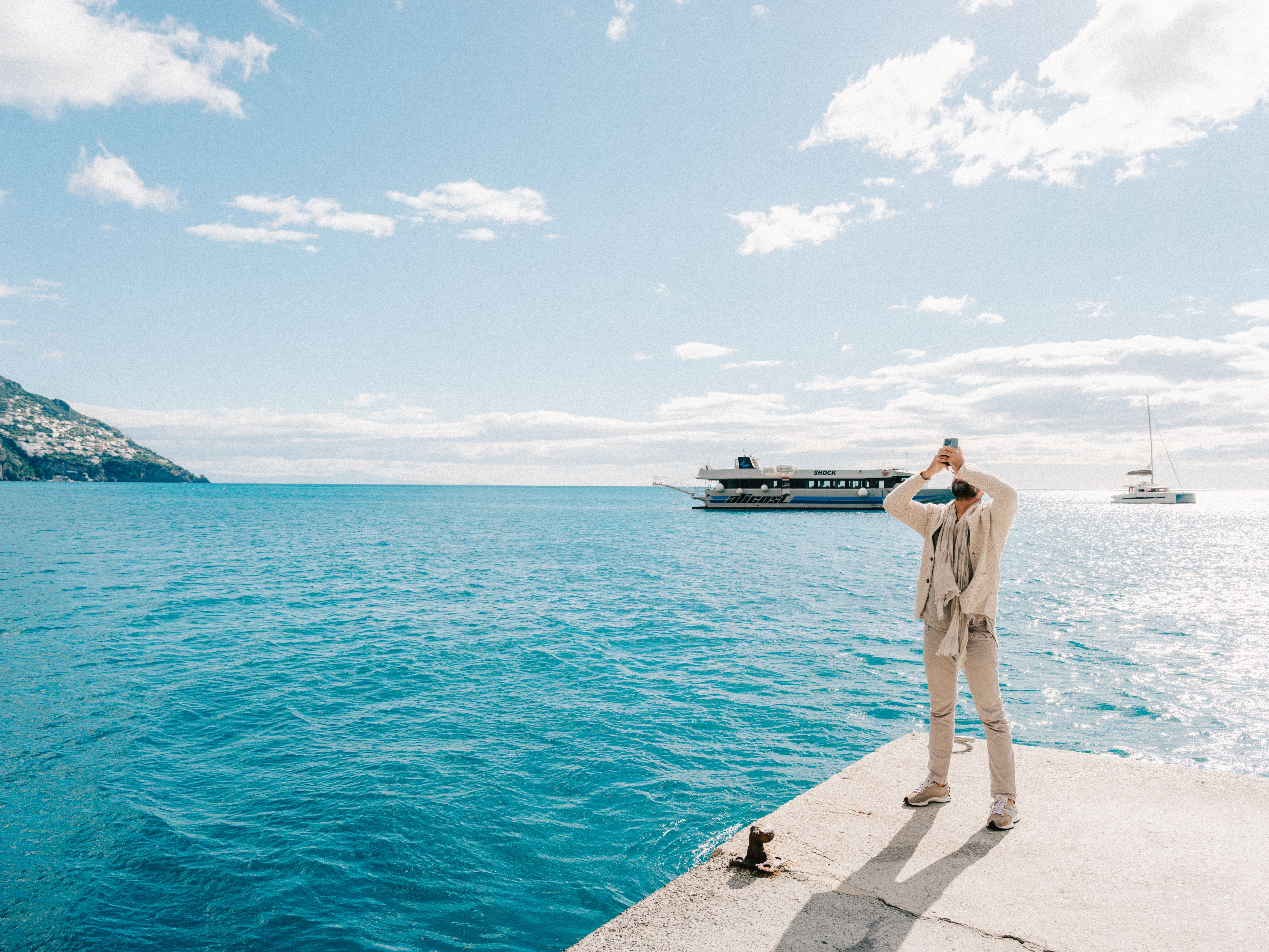 A man stands at a sunny seaside capturing the ocean view with ships in the distance.