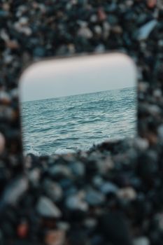 Artistic capture of the sea reflected in a mirror surrounded by pebbles on the beach, offering a unique coastal perspective.