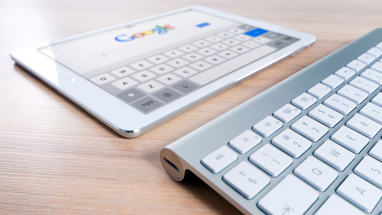 A modern tablet displaying a search engine logo next to a wireless keyboard on a wooden desk.