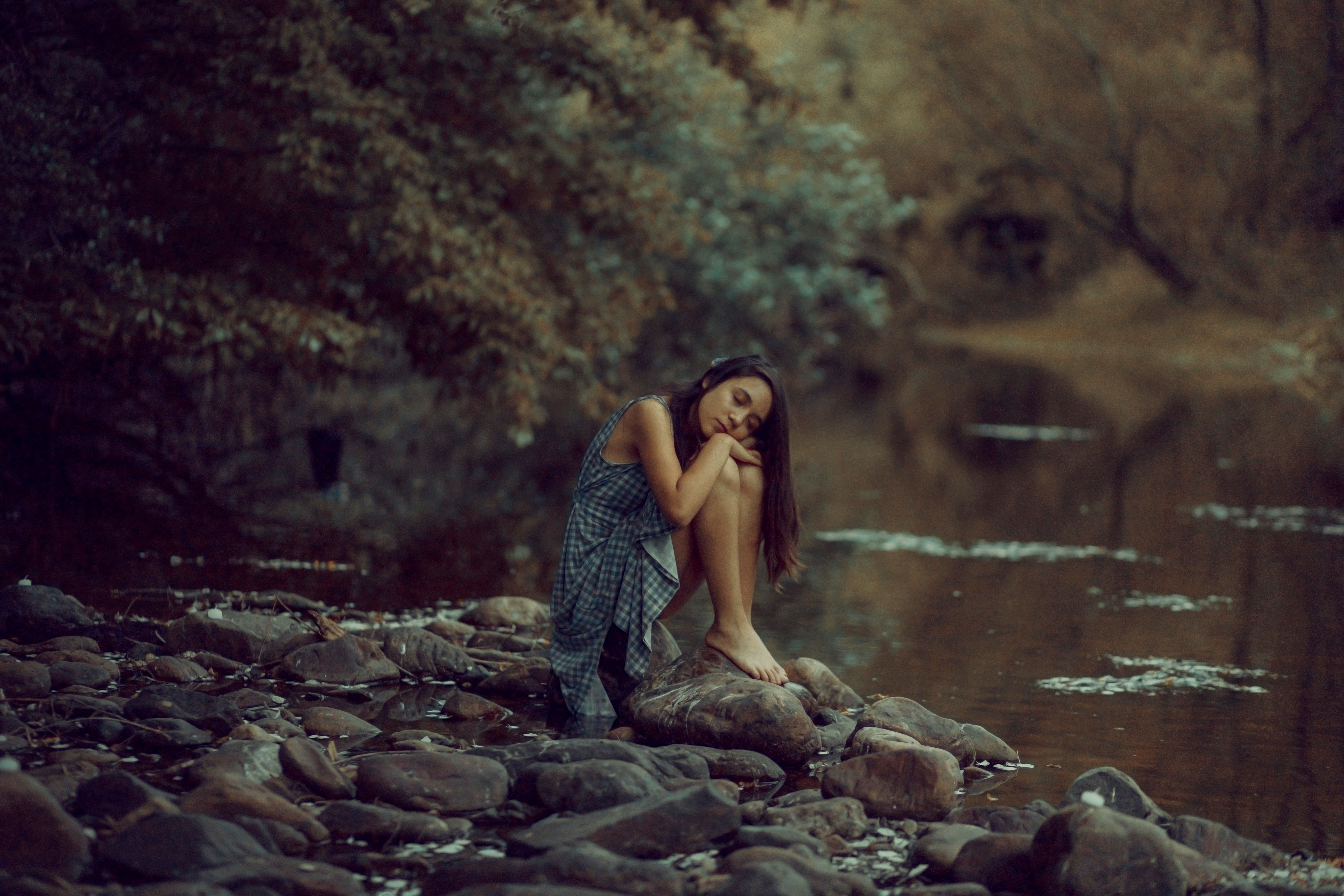 Woman sits peacefully on rocks by a serene riverbank in a forest.