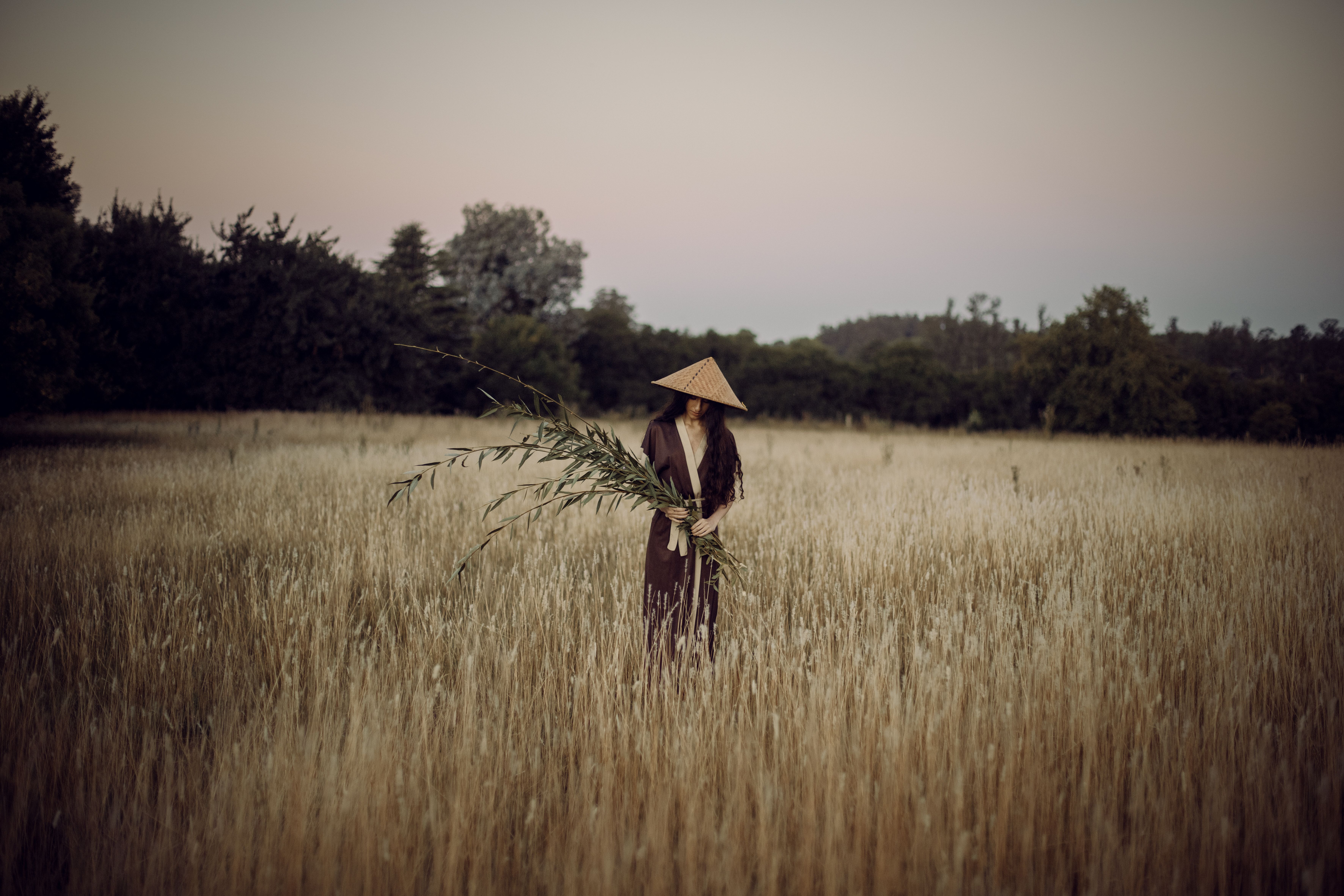 A woman in a conical hat stands amidst a serene wheat field, holding a large plant branch.