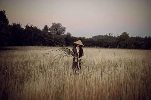 A woman in a conical hat stands amidst a serene wheat field, holding a large plant branch.
