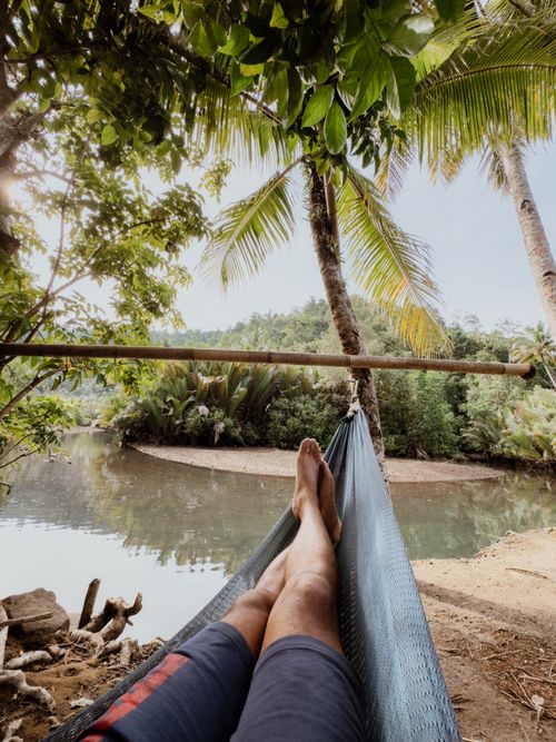 Free A man relaxes in a hammock near a tropical river surrounded by lush palm trees. Stock Photo