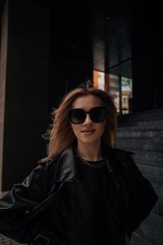 Confident woman wearing sunglasses and leather jacket with urban backdrop.