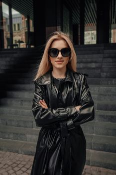 Chic model in black leather attire and sunglasses standing confidently in urban setting.
