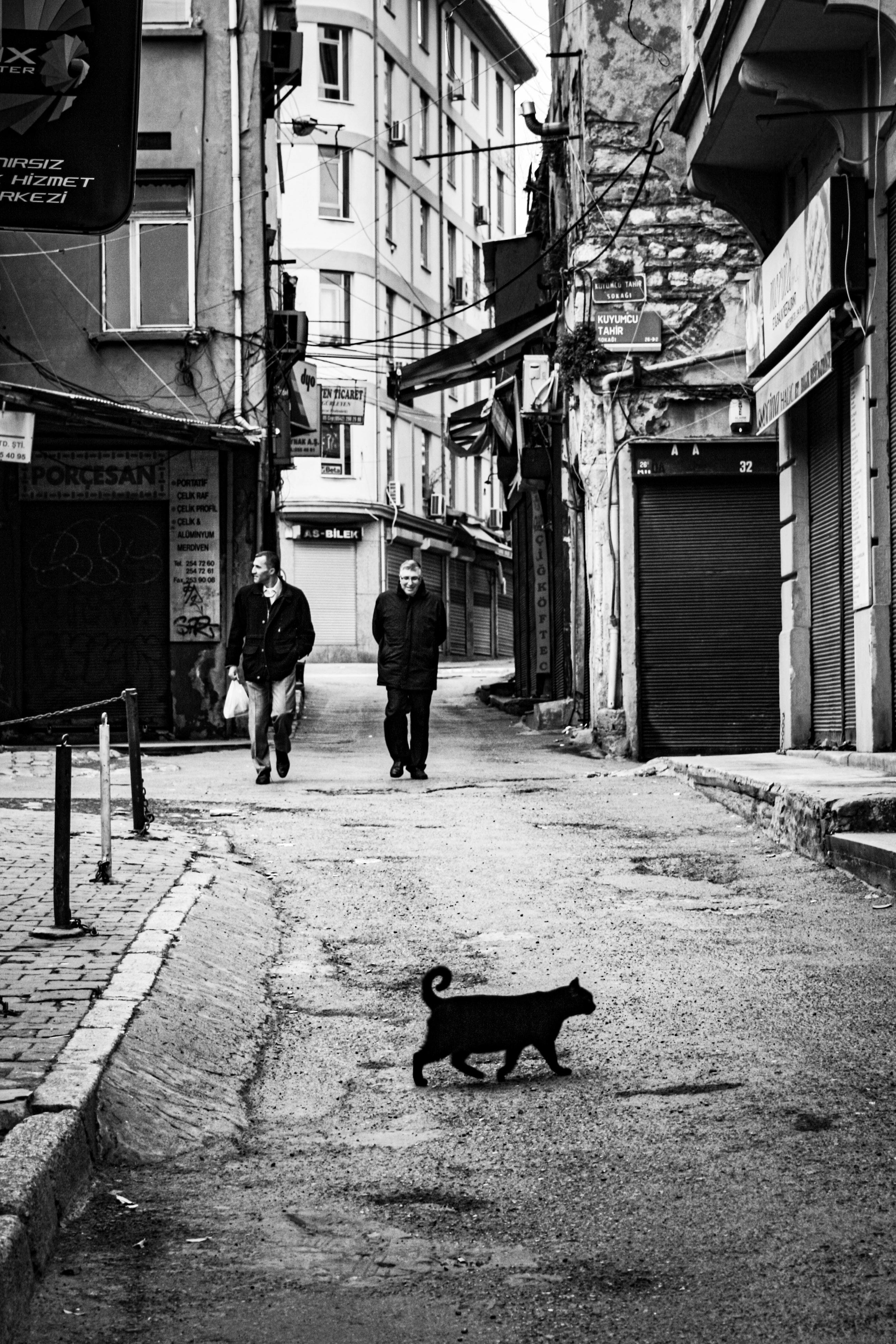 Black cat crosses an alley in Istanbul with people walking in the background, captured in monochrome.