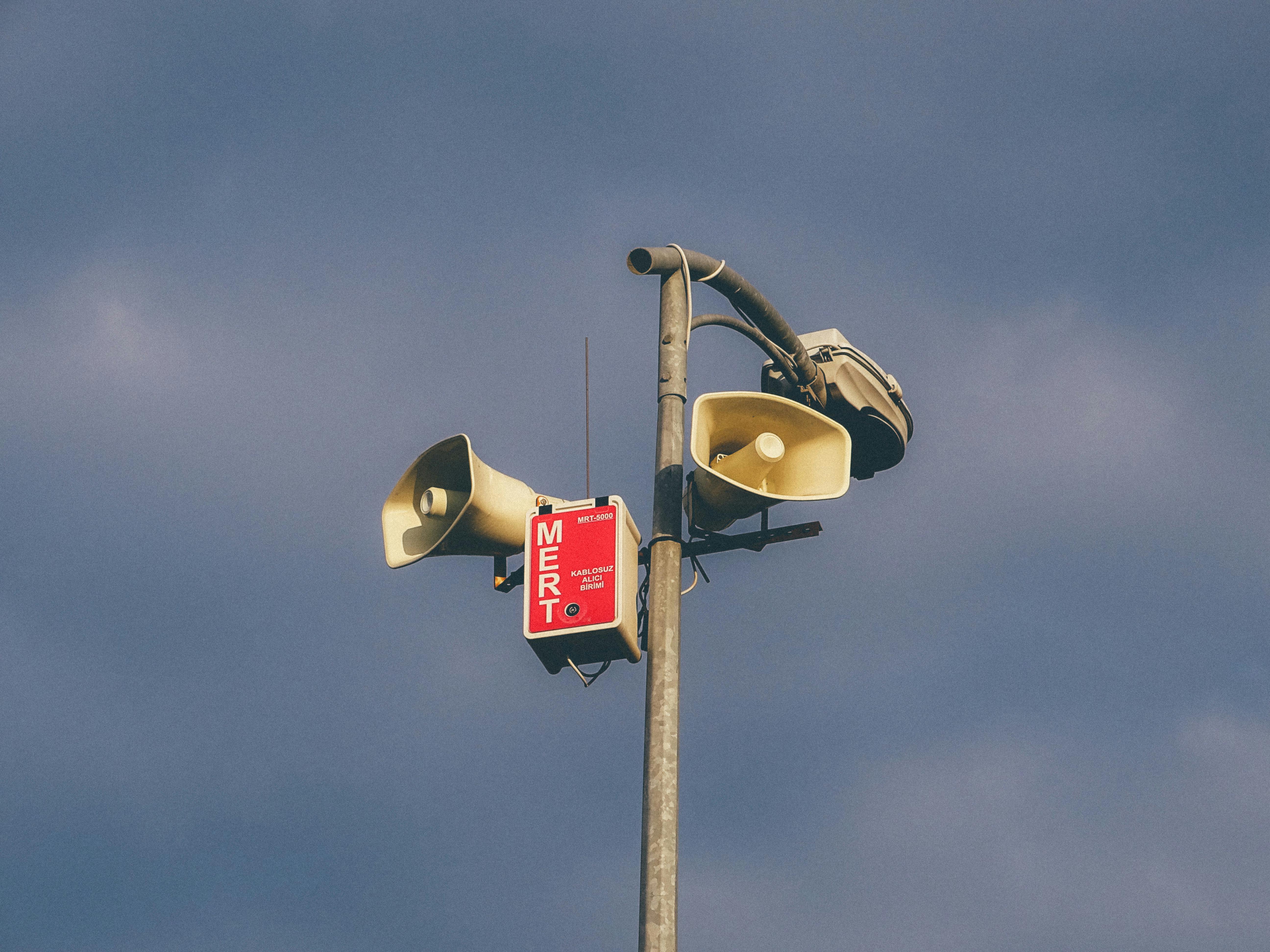 A warning system with loudspeakers on a pole against a cloudy sky, signaling emergency alerts.