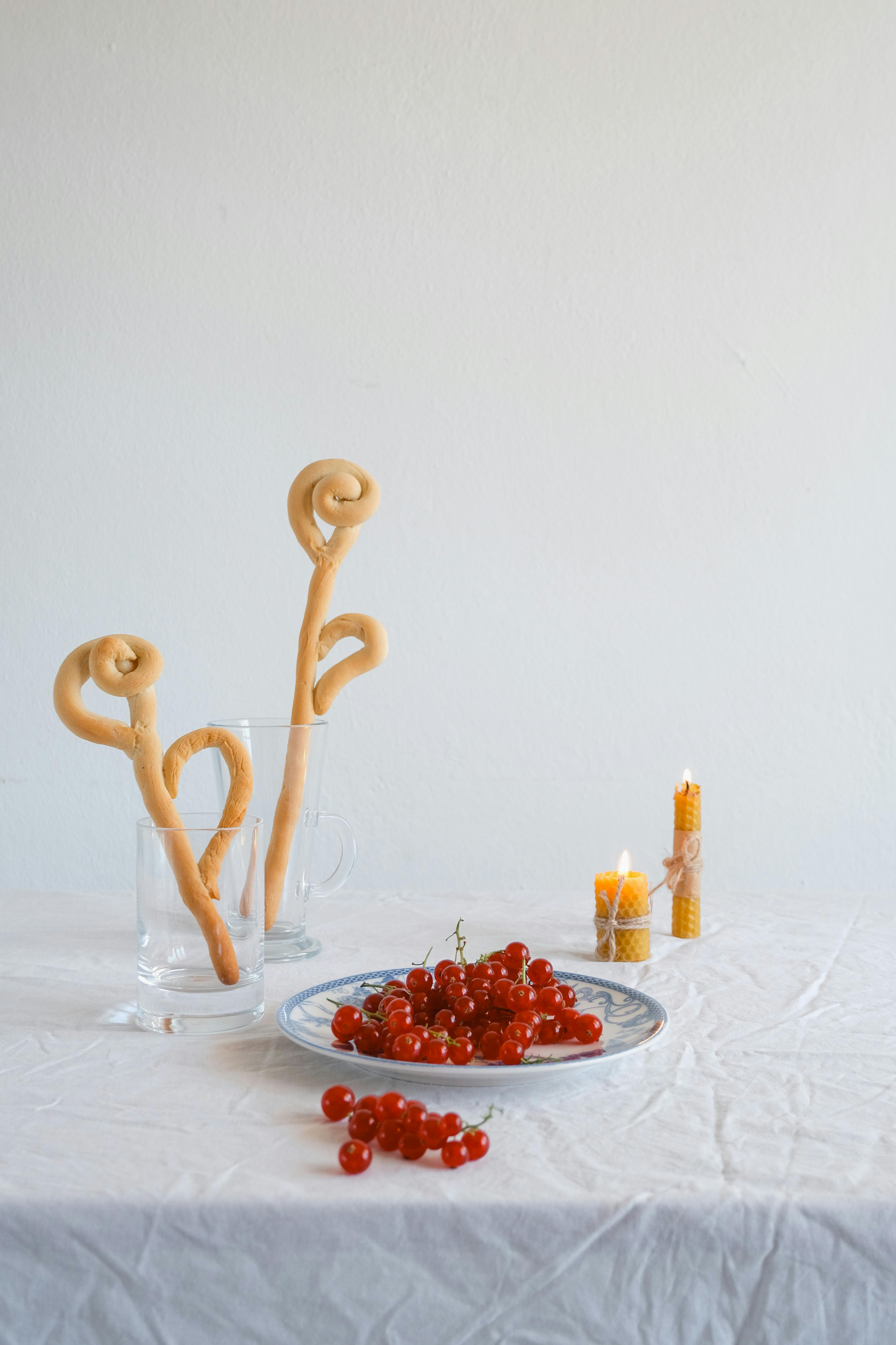 Elegant still life featuring dough shapes in glasses, red currants on a plate, and lit candles for a cozy ambiance.