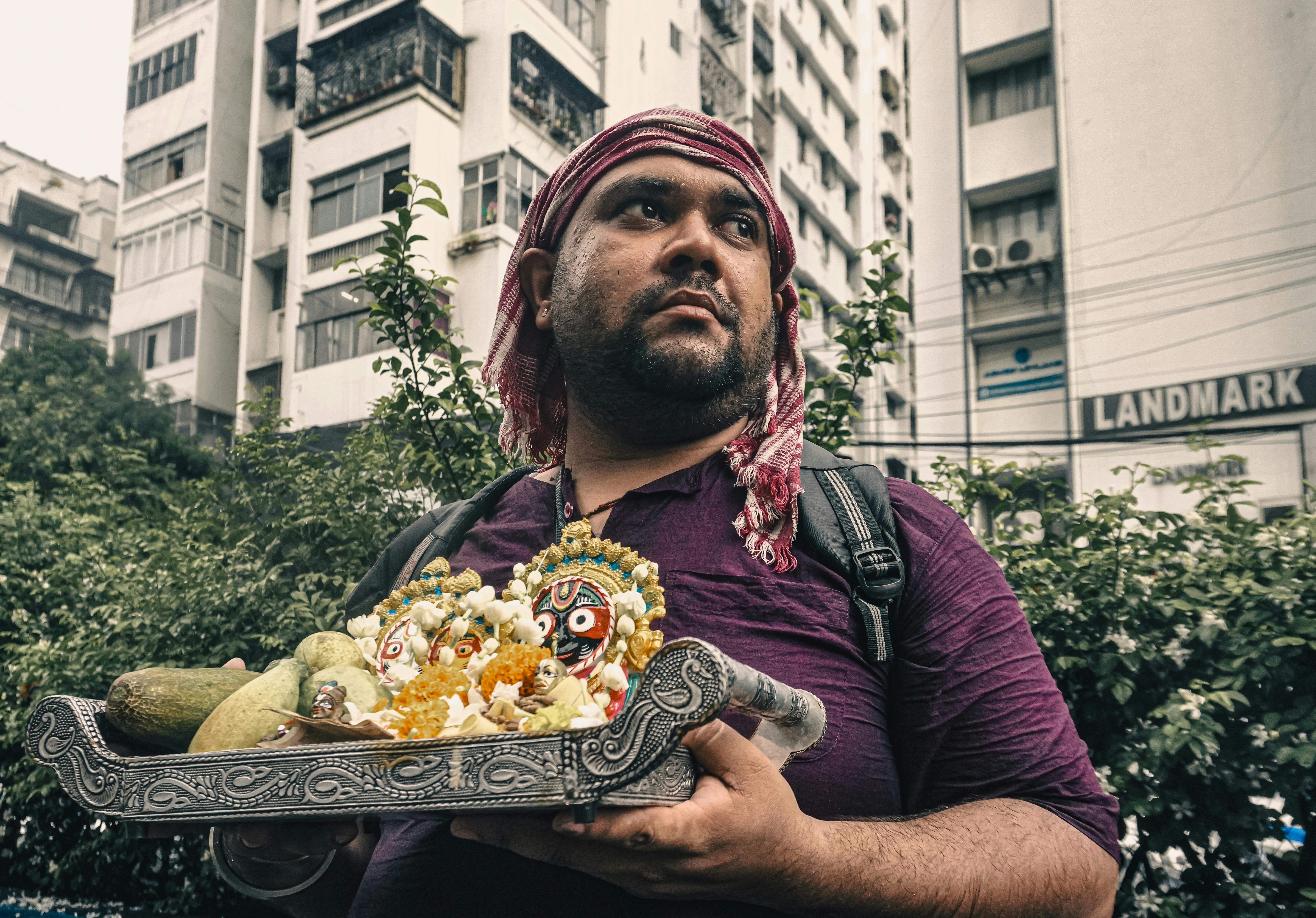 Man with headscarf holds a decorated tray with Hindu deities in a city backdrop.