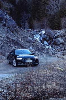 Black Audi car driving through scenic Romanian countryside with waterfall and forest backdrop.