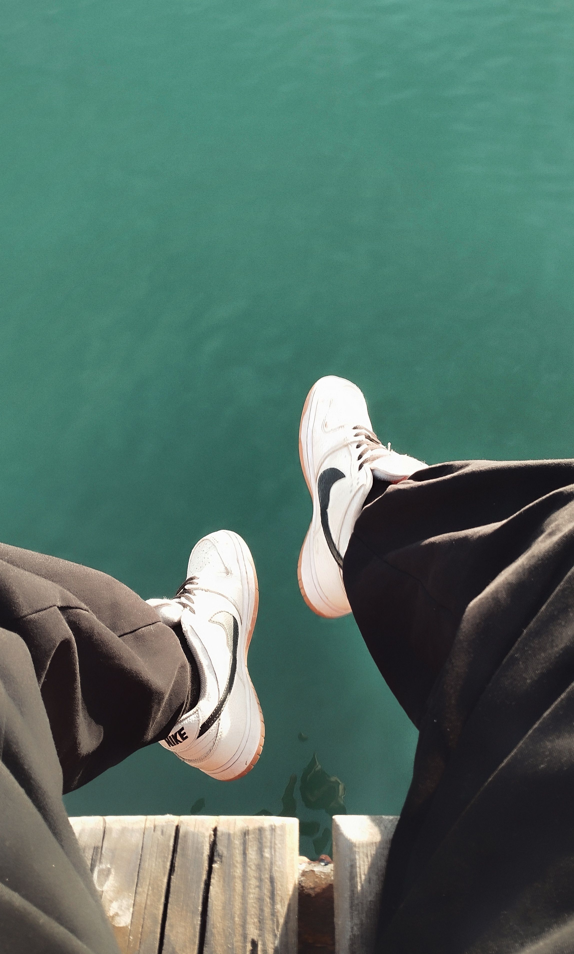 Legs hanging over a wooden boardwalk by water, featuring white sneakers for a leisurely vibe.