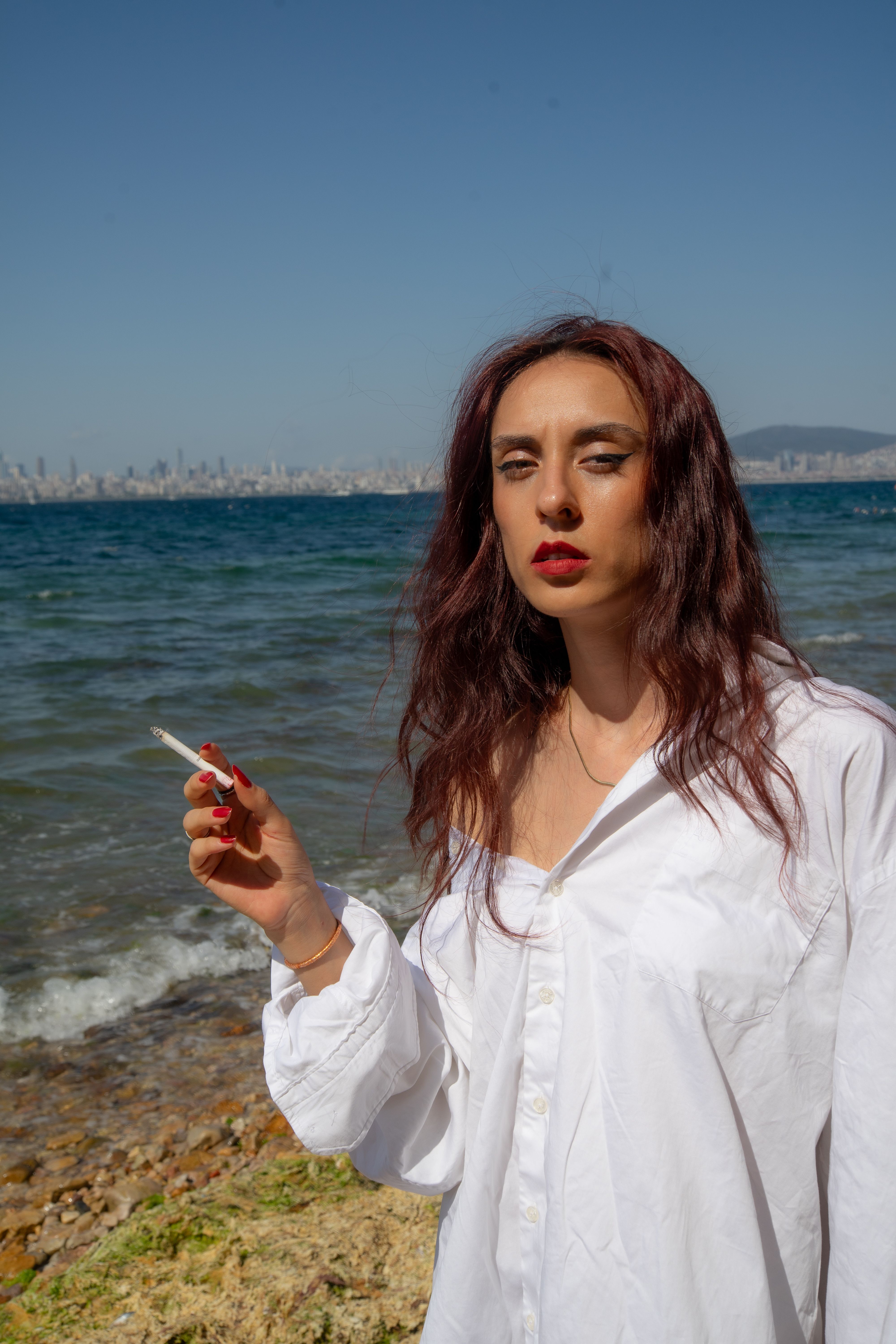 A woman stands by the ocean holding a cigarette, exuding a sense of mystery and style.