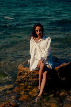 Woman sitting on a rocky seashore in summer, wearing a white shirt with a serene expression.