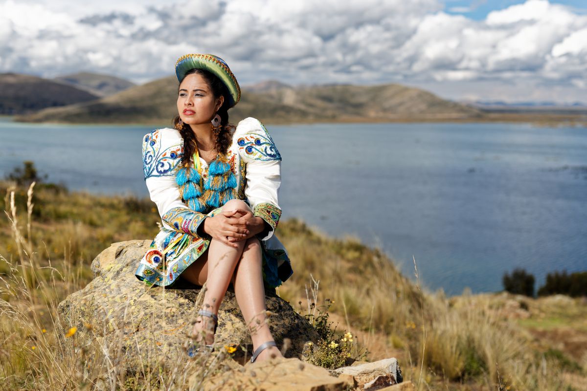 A Bolivian woman in a pollera skirt and embroidered shawl sitting on the shore of Lake Titicaca under a bright sky