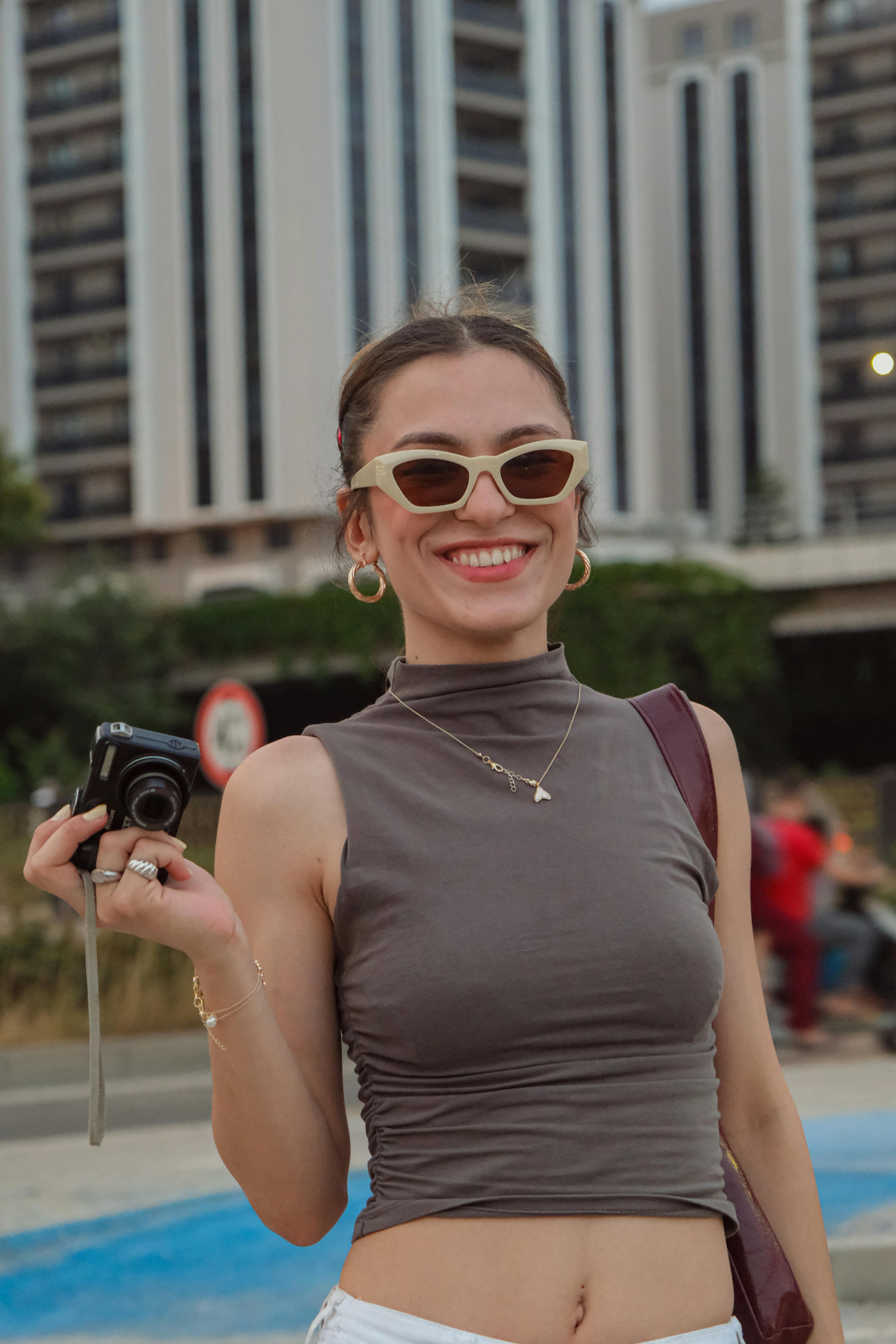 Fashionable woman smiles while holding a camera in a vibrant city environment.