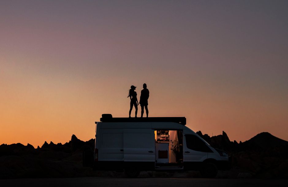 A couple stands silhouetted on a van's rooftop against a colorful sunset sky.