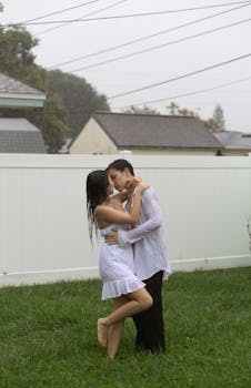 A young couple shares a passionate kiss in the rain, standing in a lush garden with a white fence backdrop.