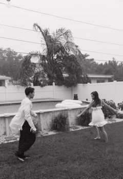A couple enjoys a playful dance in the rain on a lush lawn with a poolside backdrop.