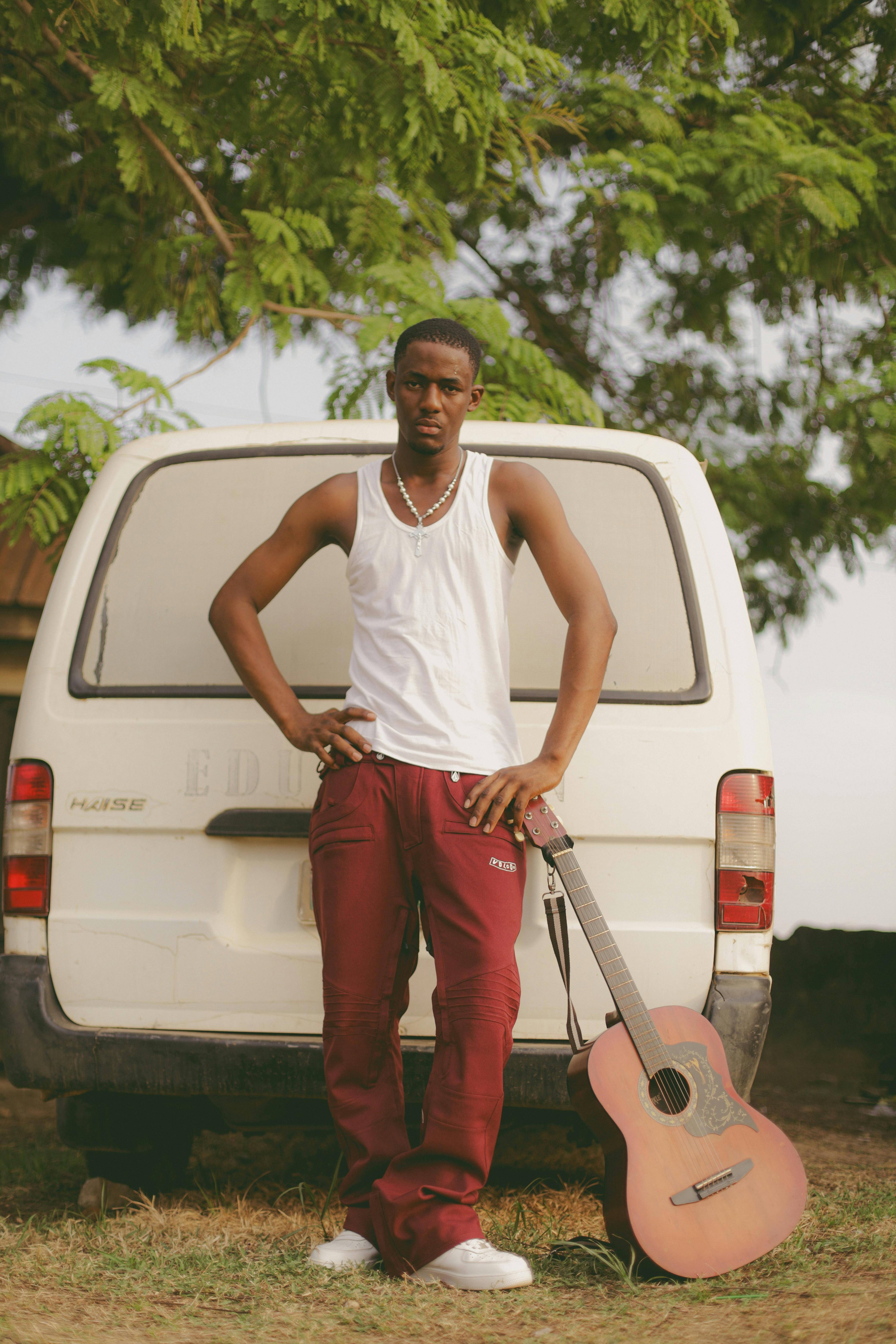 A young Nigerian man poses with a guitar against a van in an outdoor setting.