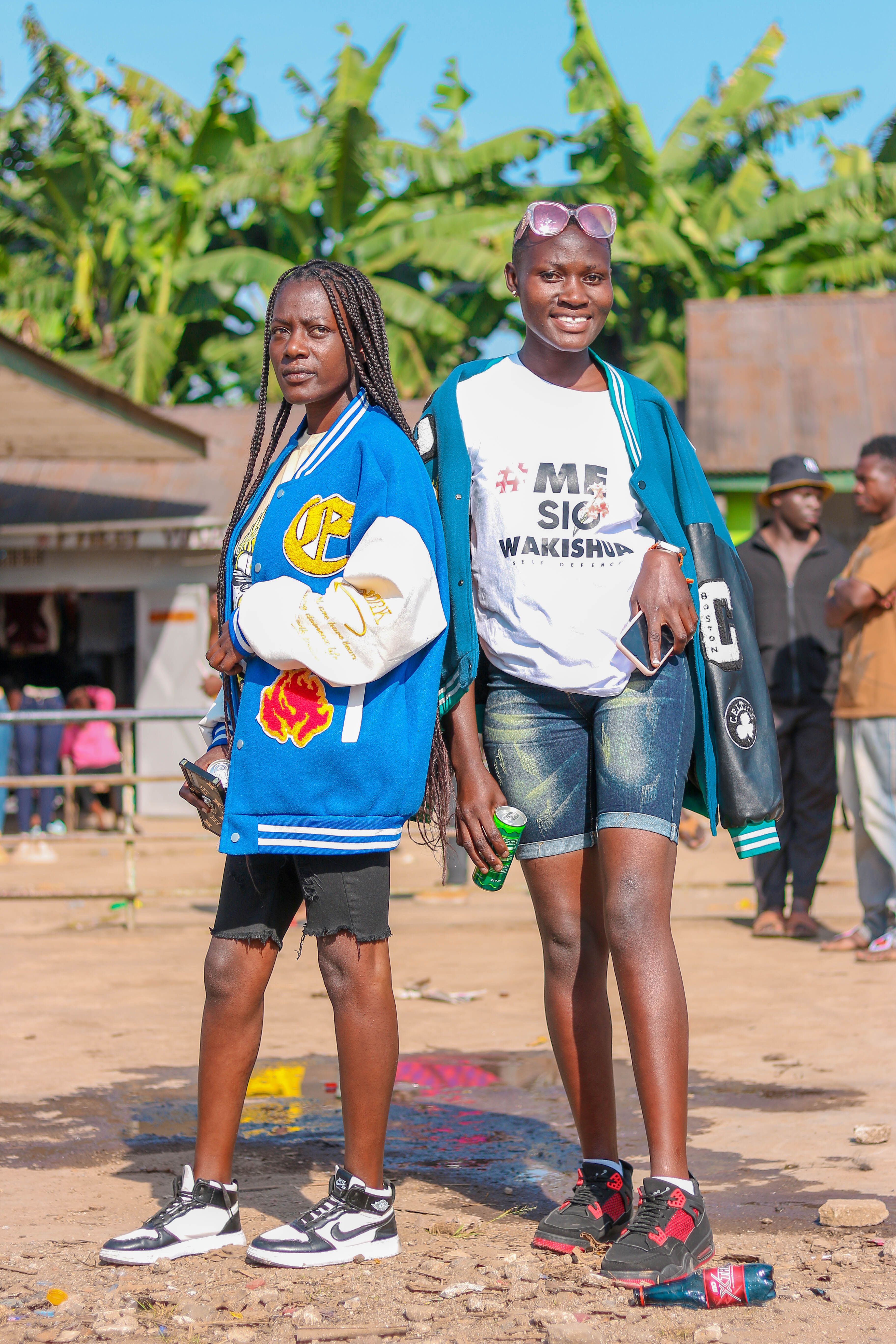 Two young women in varsity jackets enjoying a sunny day at an outdoor market.