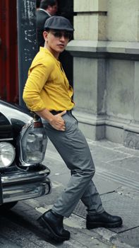 Fashionable man in a yellow shirt and cap leaning on a vintage car in an urban environment.