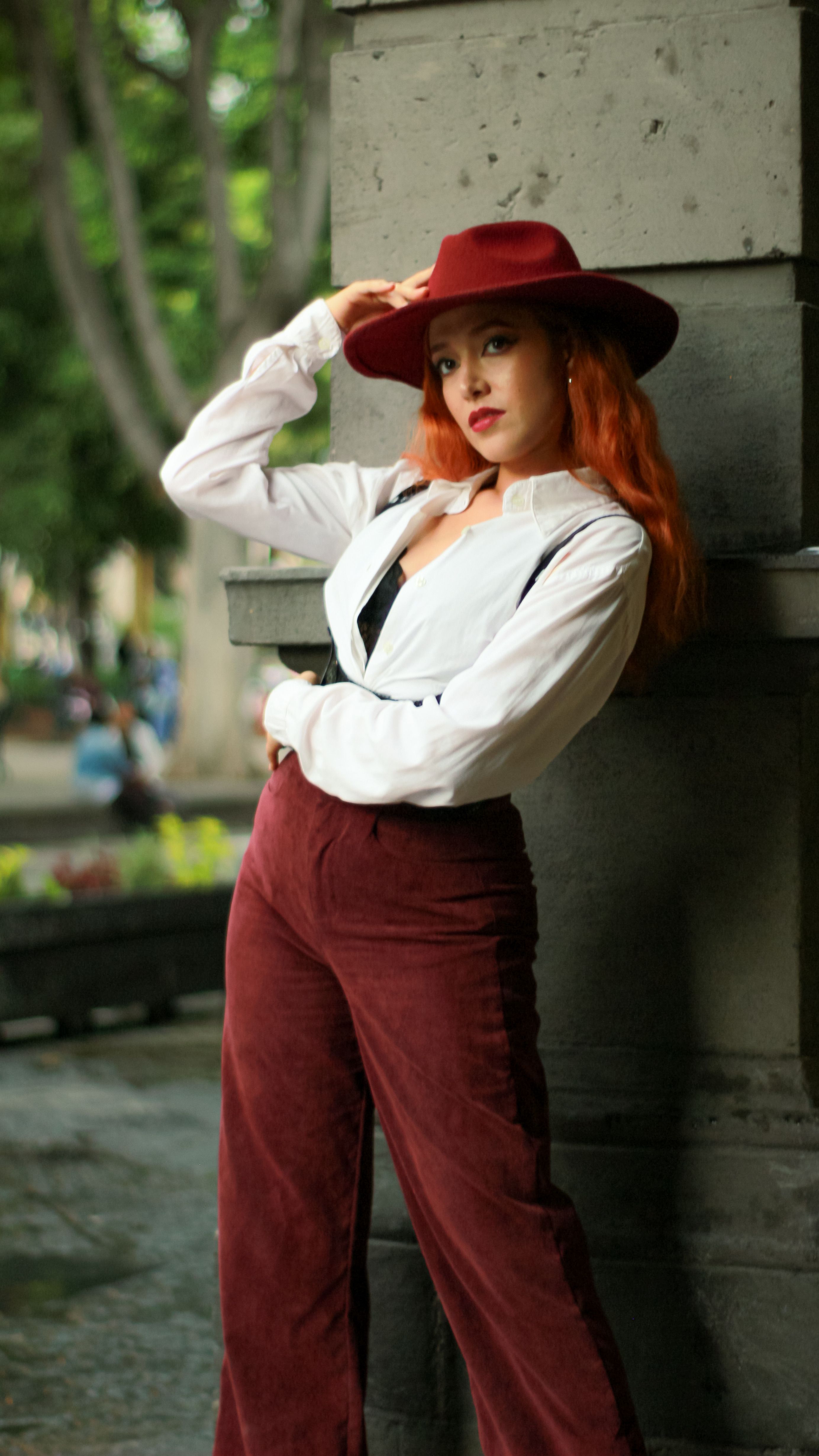 Mulher estilosa com cabelo vermelho posa em um look chique com um chapéu vermelho e calças de veludo cotele ao ar livre.