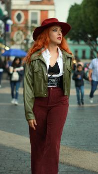 Stylish woman with red hair walking in urban environment, wearing hat and burgundy pants.