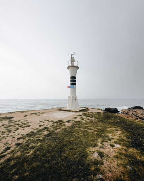 Free A solitary lighthouse stands on a rocky shore overlooking a calm ocean under a hazy sky. Stock Photo