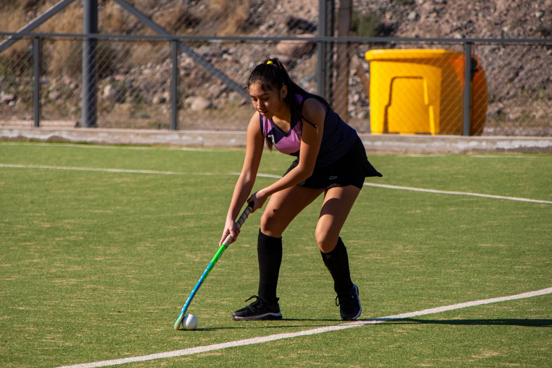 Woman playing field hockey on a sunny day, showcasing determination and skill.