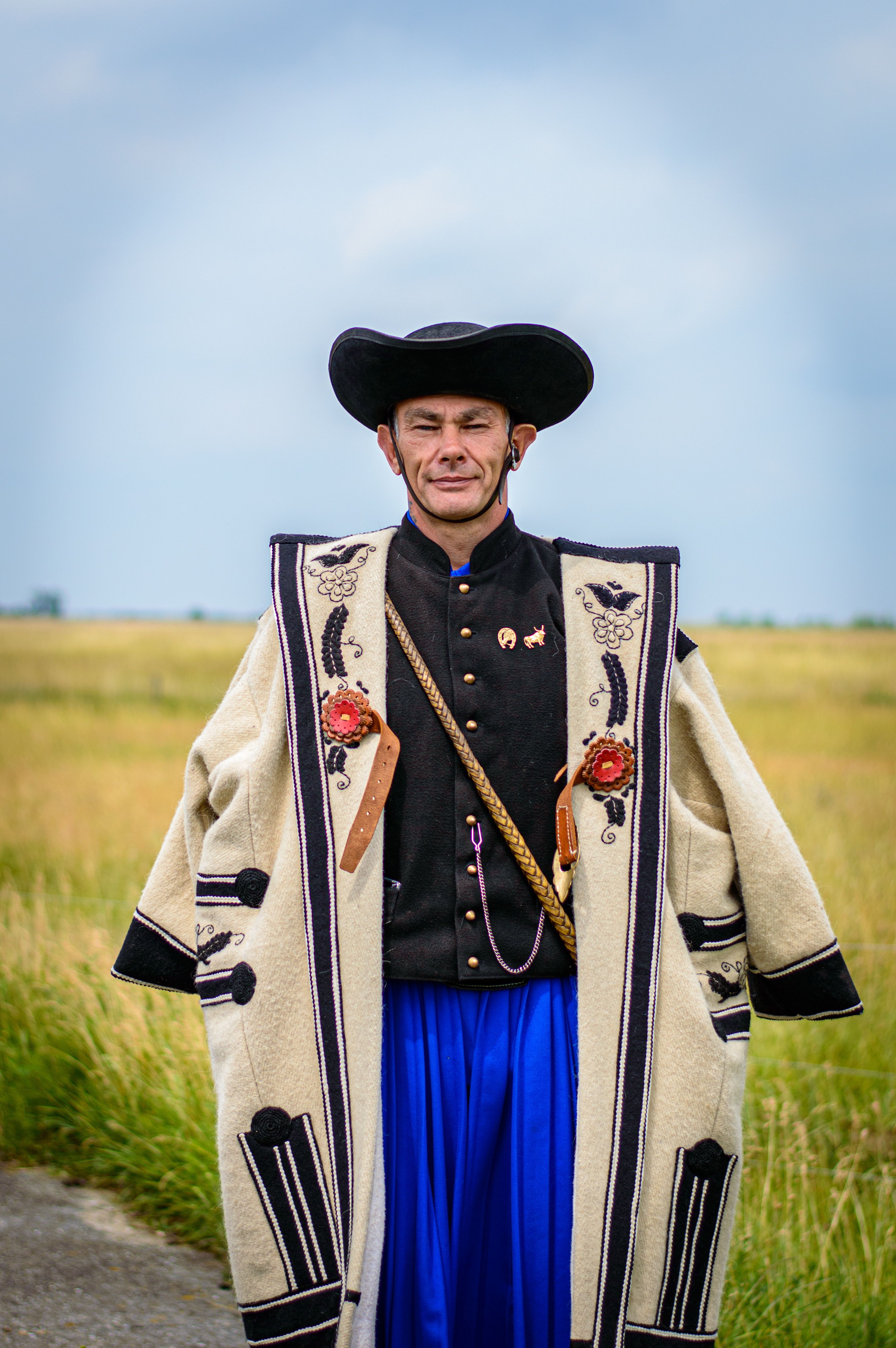 Portrait of a man in a traditional embroidered coat and hat, posing outdoors in a rural field.