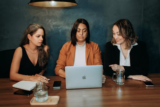 Three women engaged in a work discussion around a laptop in a professional setting.