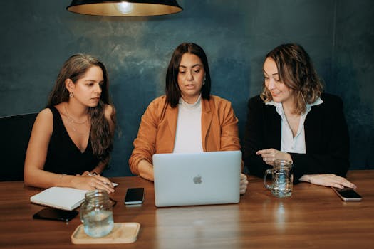 Three women working at a wooden table with a laptop in a modern office environment.