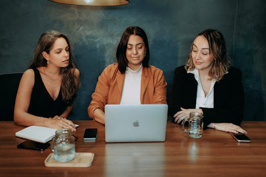 Three women engaged in a focused business meeting at a modern office setting with laptops and notebooks.