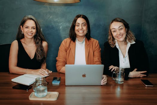 Three professional women collaborating at a desk with a laptop, notebooks, and phones, indoors.