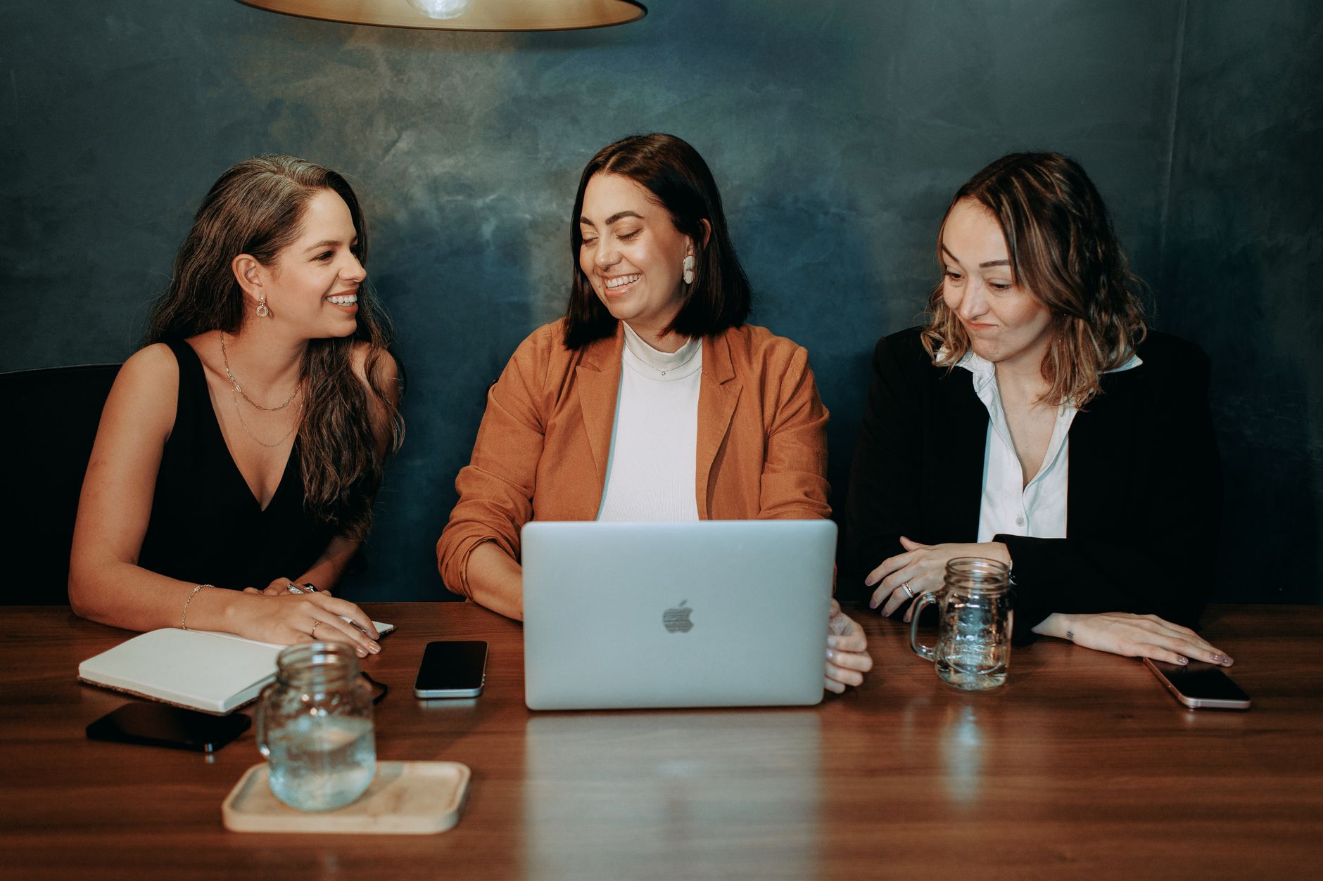 Three women collaborating in a modern office setting with laptops and notebooks.