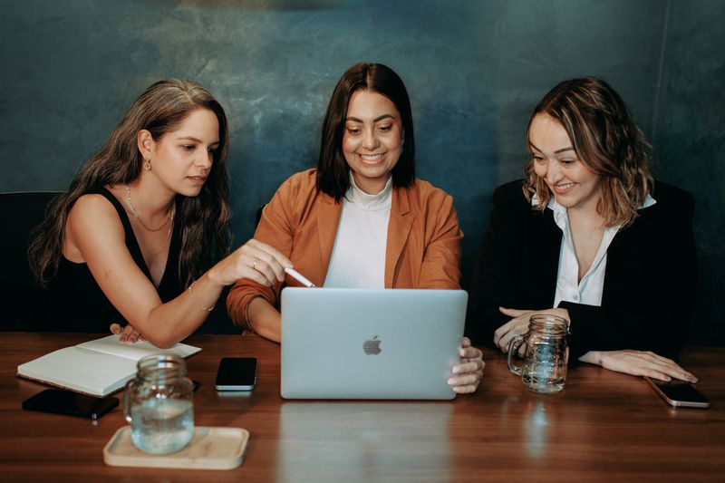 Three women engaged in a business meeting, collaborating on a laptop in a modern office setting.