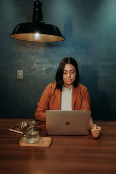 A woman in a brown blazer intently uses a laptop at a wooden table with soft lighting.