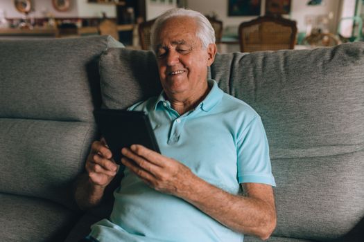 Smiling elderly man using a tablet while sitting comfortably on a sofa, embracing modern technology.