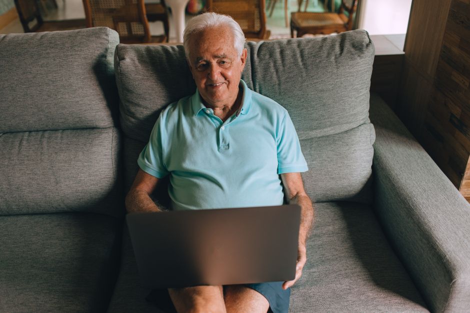 Senior man enjoying laptop usage on a comfy sofa indoors, showcasing technology engagement.
