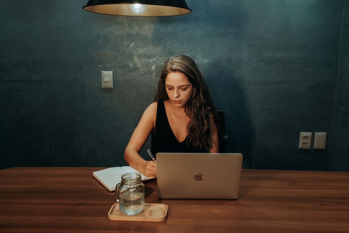 Someone writing in a notebook next to a laptop at a desk