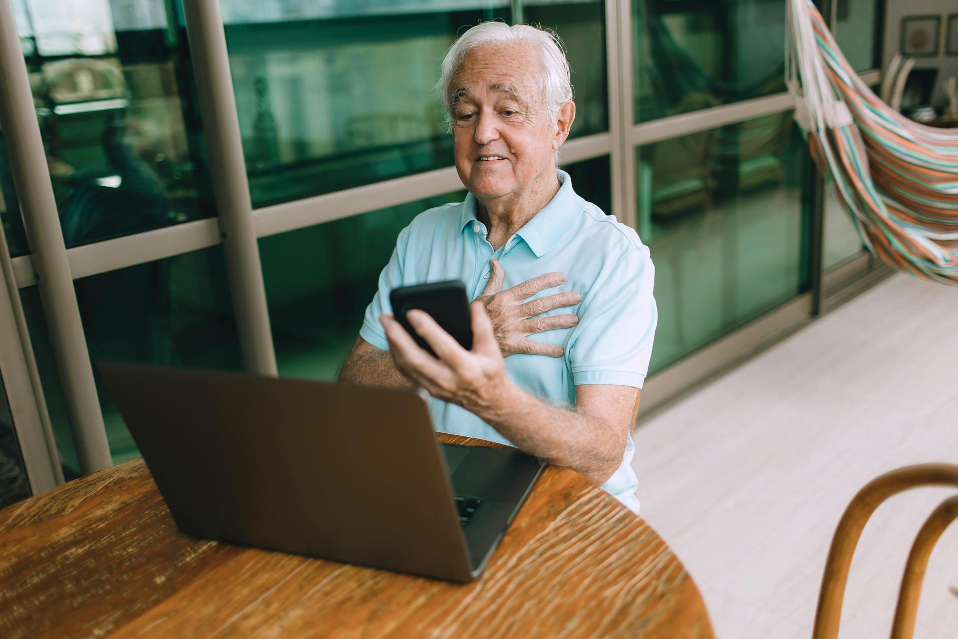 Senior man enjoying video chat with smartphone and laptop indoors. Modern technology and communication concept.